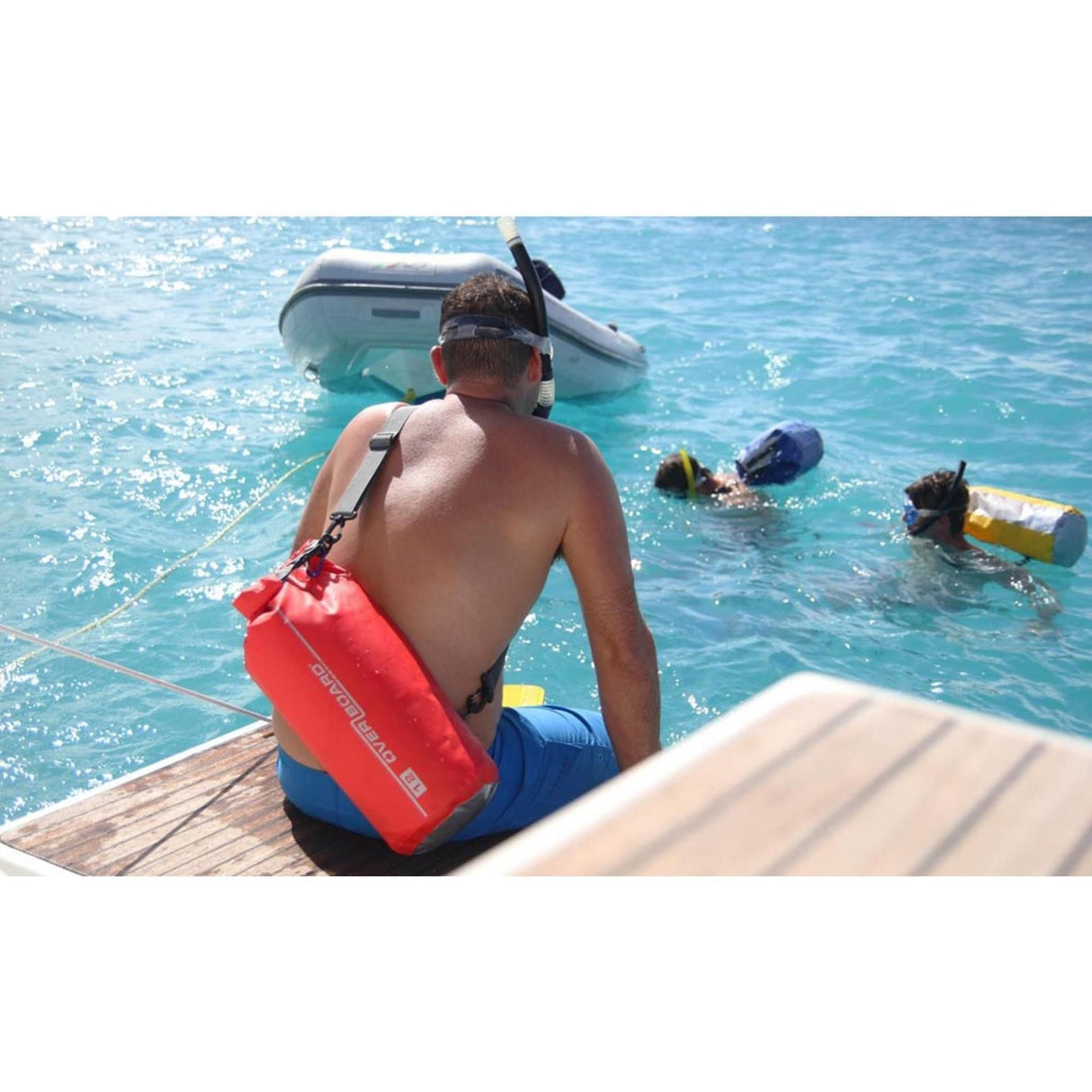 OverBoard wasserdichter Packsack 12 Liter rot, Mann sitzt am Steg mit rotem Dry Bag, Wasser und Boot im Hintergrund