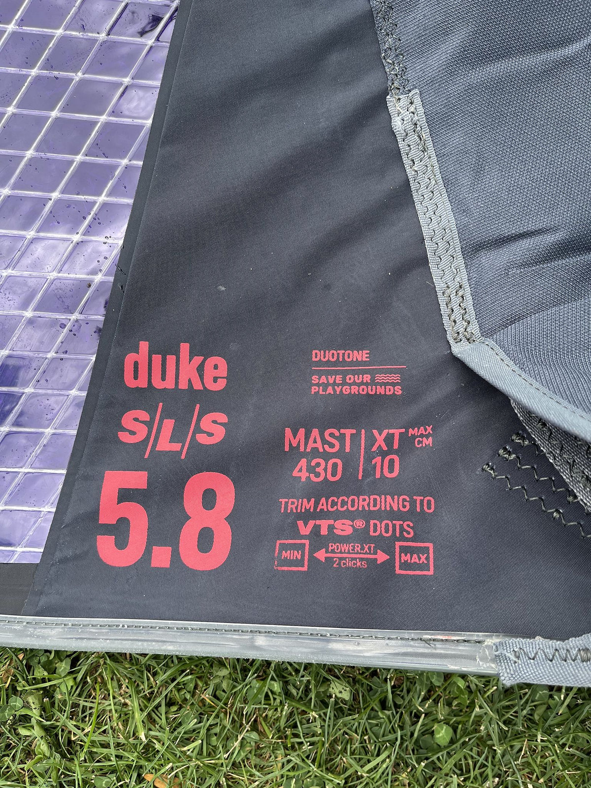 Duotone Duke SLS 5.8 Kite 2026, grau mit rot-violettem Duotone-Design, Detailansicht des Segels mit technischen Spezifikationen und Markenlogos auf Gras liegend