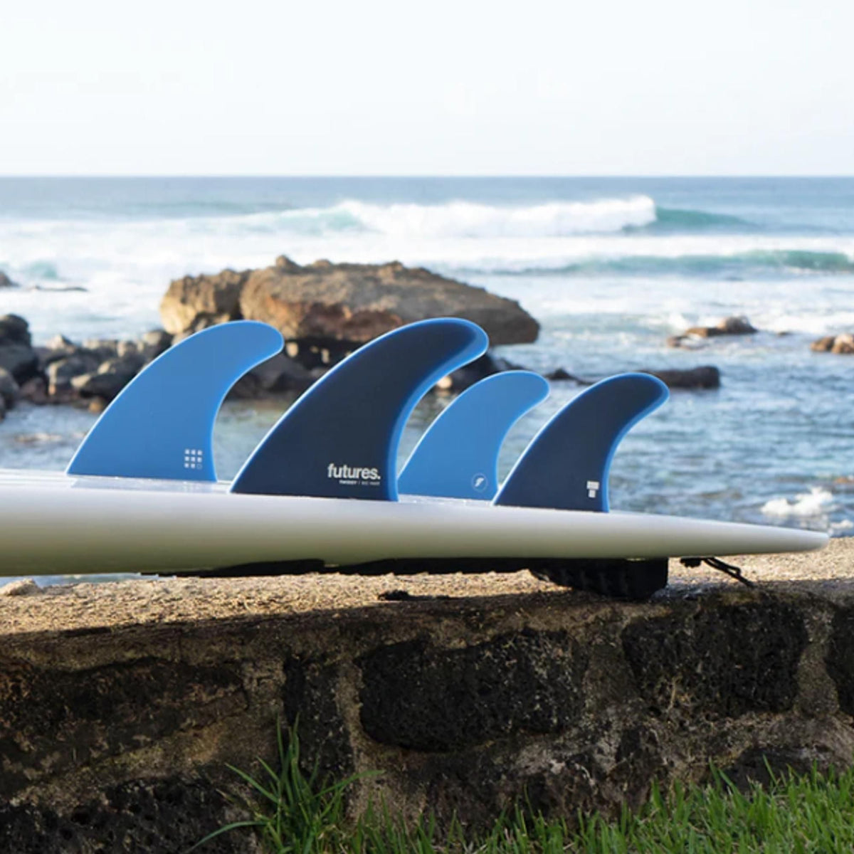 Futures Twiggy Big Wave 5-Fin Set, vier blaue und schwarze Surfflossen aus Kunststoff auf weißem Surfboard-Rail am Strand