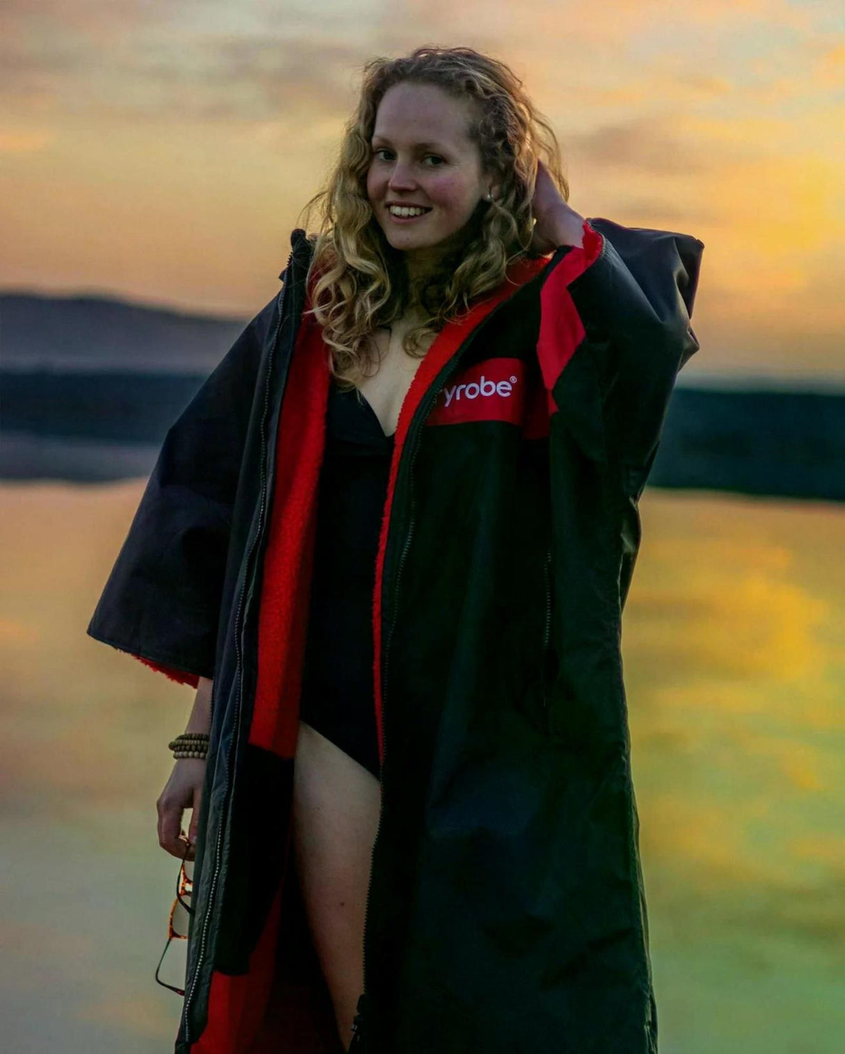 DRYROBE Advance Kurzarm in Marineblau und Rot, getragen von Person mit Surfbrett am Strand bei Sonnenuntergang