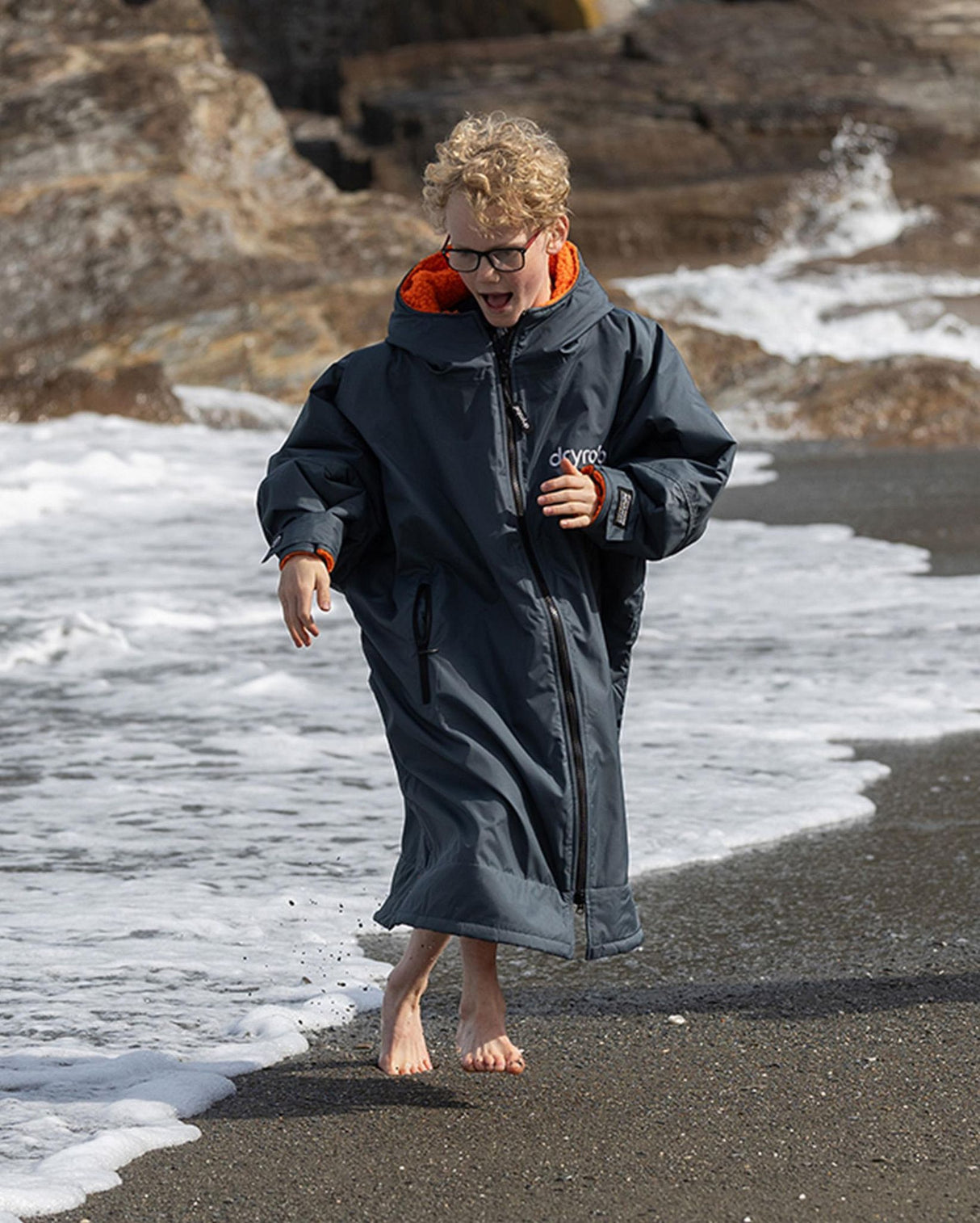Dryrobe Advance Long Sleeve Kids in Marineblau mit orangefarbenem Kragen, Kind läuft barfuß am Strand