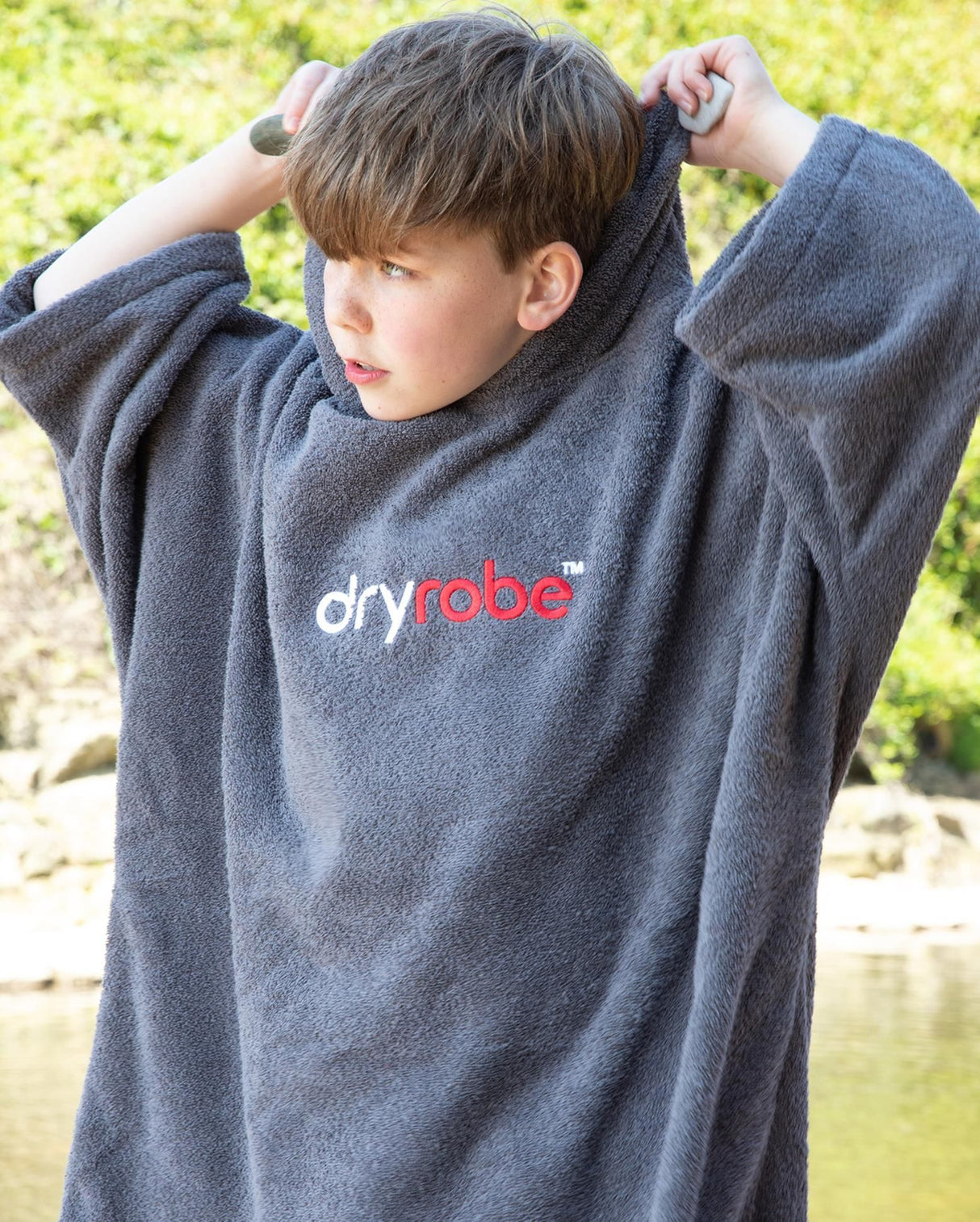 Dryrobe Handtuchmantel Bio Kids in Grau, Kind trägt Mantel mit Kapuze und Dryrobe-Logo, Frontalansicht im Freien