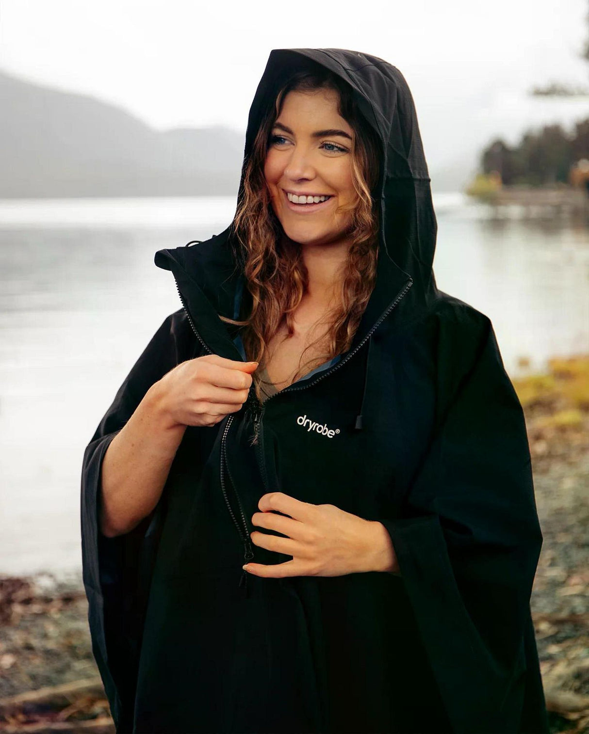 Dryrobe Regen-Poncho in Schwarz, wasserdicht, mit Kapuze, getragen von einer Frau am See