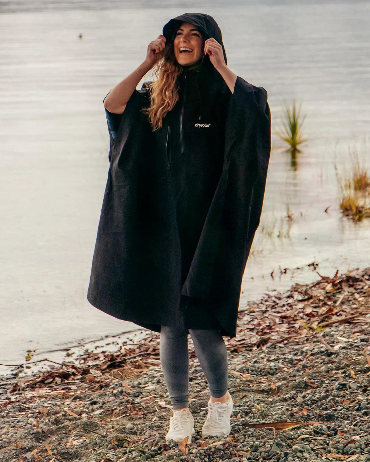 Dryrobe Regen-Poncho in Schwarz, wasserdicht, Frau trägt Poncho mit Kapuze am Strand, Frontalansicht