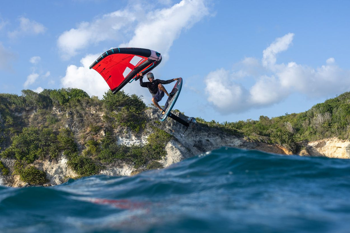 Duotone Unit 2027 Kiteboard mit rotem und weißem Segel über Meerwellen, Fahrer in Aktion, Küstenlandschaft im Hintergrund