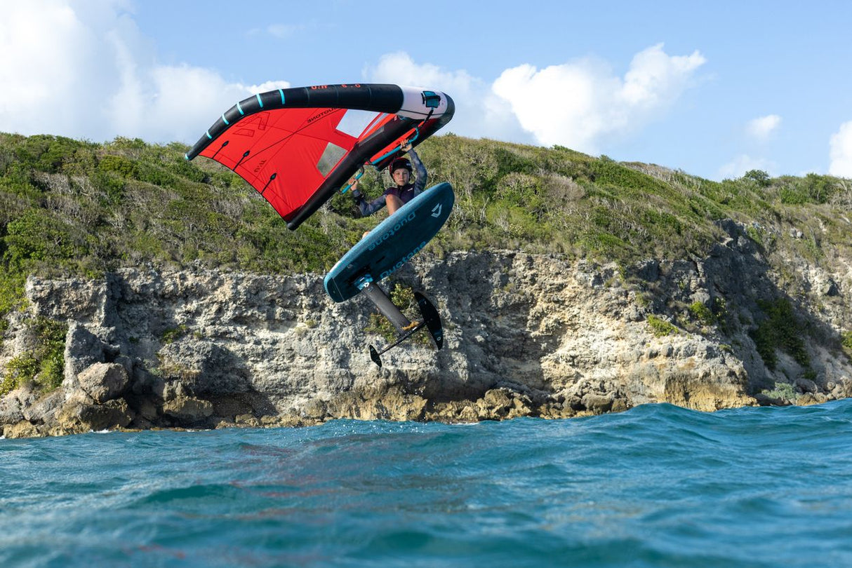 Duotone Unit 2027 Kiteboard rot-dunkelblau in Aktion über Wasser vor Felsenküste