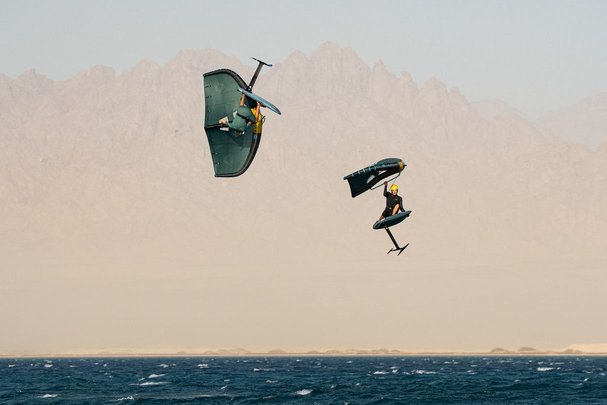 Duotone Unit SLS 2027 Kiteboard grün-schwarz in Aktion über Wasser mit Kitesurfer, Frontalansicht gegen Bergkulisse