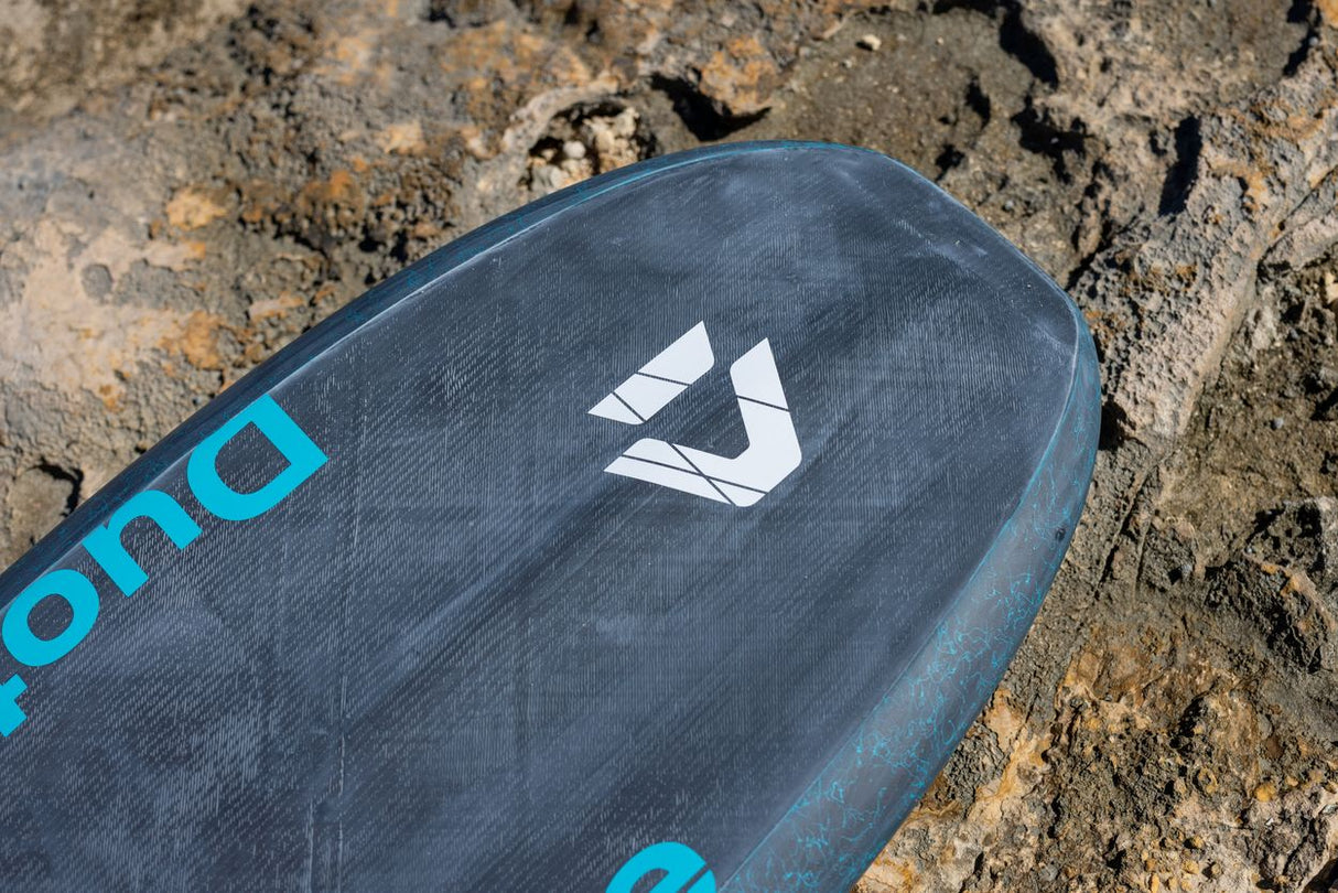 Duotone Sky Style SLS 2027 Kiteboard mit dunkelblauem Deck, weißem Logo und türkisen Beschriftungen im Sand