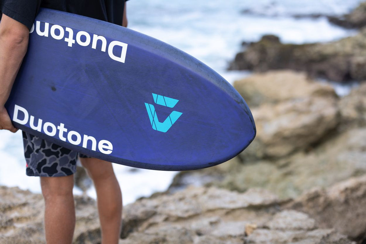 Duotone Crush SLS 2027 Kiteboard in Blau mit Duotone-Logo, von Person am Strand gehalten