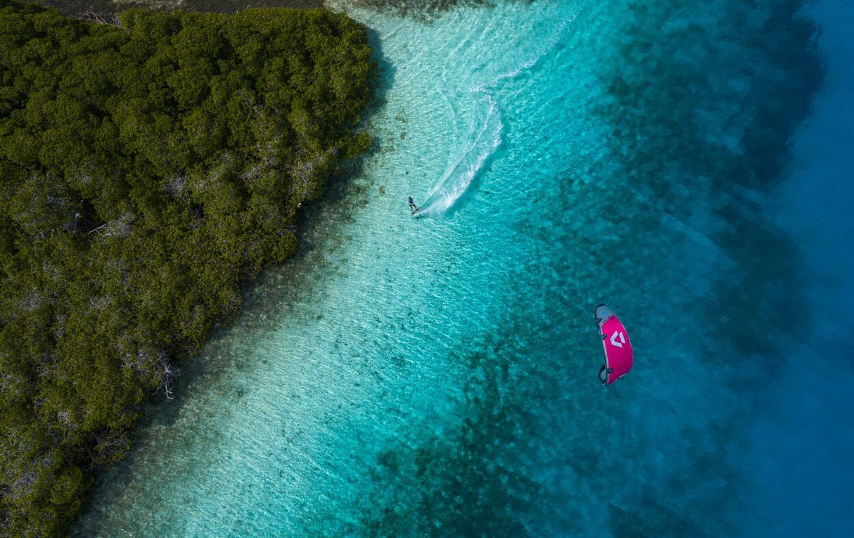 Duotone Jaime Concept Blue 2027 Kiteboard in Pink auf türkisblauem Wasser, Luftaufnahme mit Küstenwald