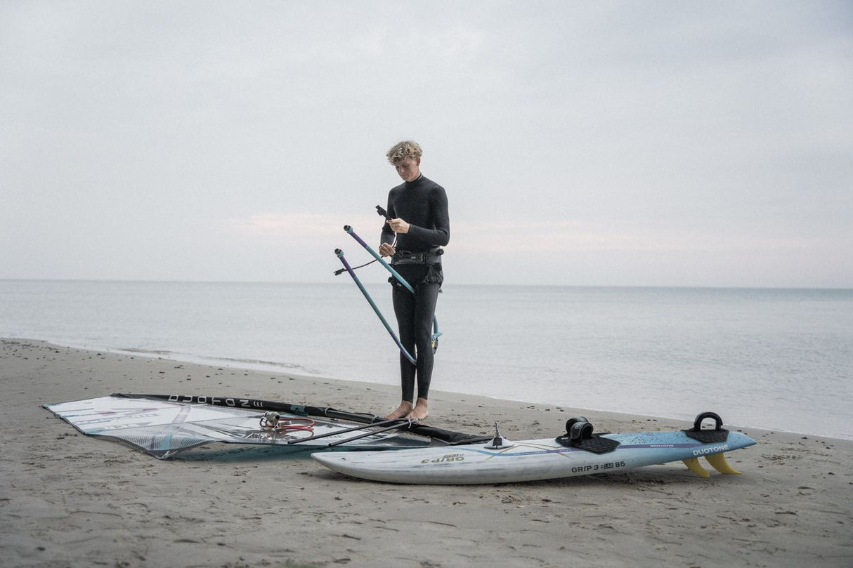 ION Harness Line 2026 schwarzes Trapez, Athlet steht mit Kiteboard am Strand, Frontalansicht, Wassersportausrüstung