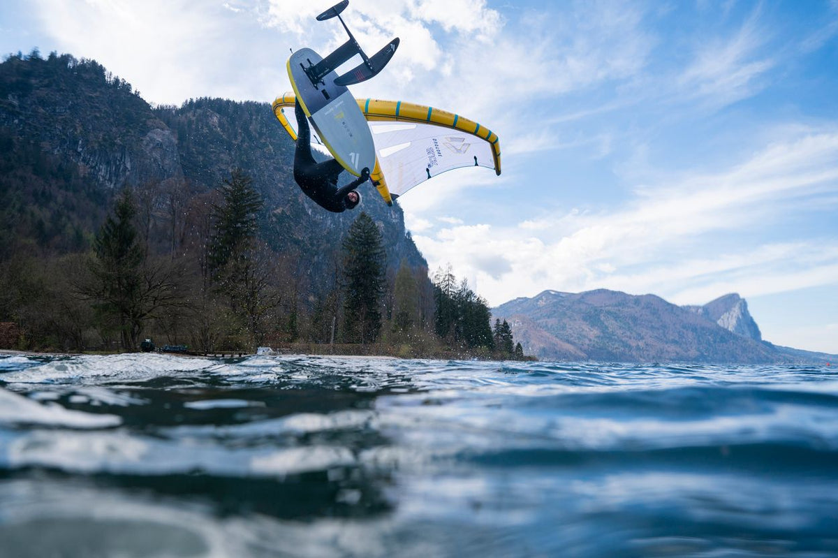 ION Hybrid Gloves 1+2.5 2026 schwarz, Kitesurfer in Aktion über Wasser mit Berg im Hintergrund