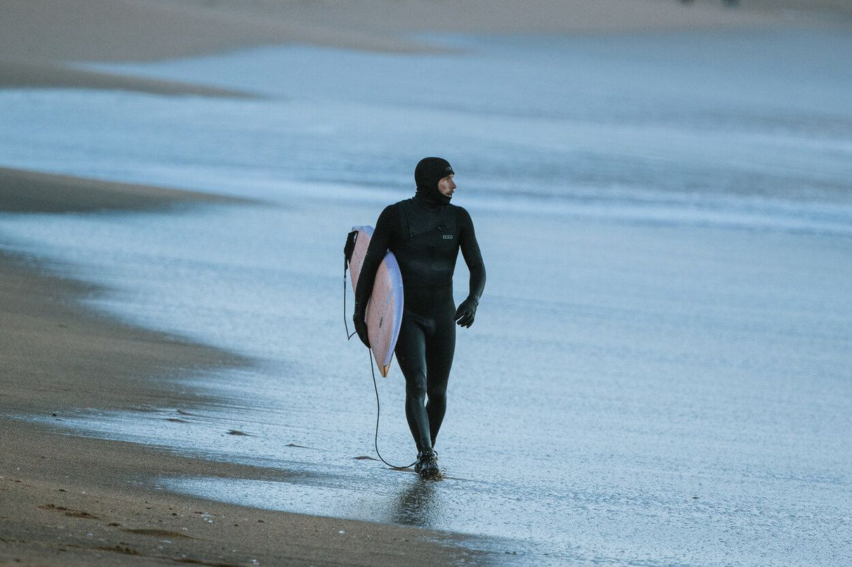 ION Ballistic Boots 3/2 Round Toe schwarz, Surfer am Strand mit Neoprenanzug und Surfboard, Ansicht von vorne