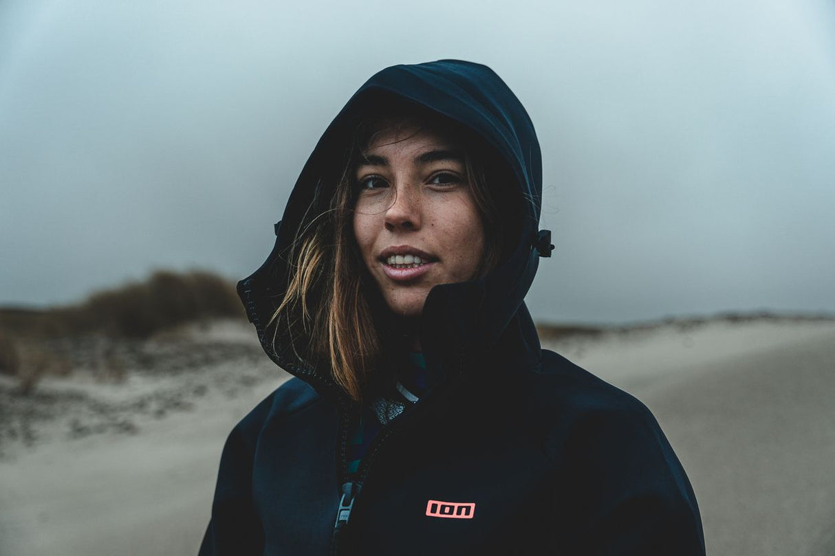 ION Neo Cosy Coat 2026 dunkelblau mit Kapuze, Frau trägt Neoprenjacke am Strand