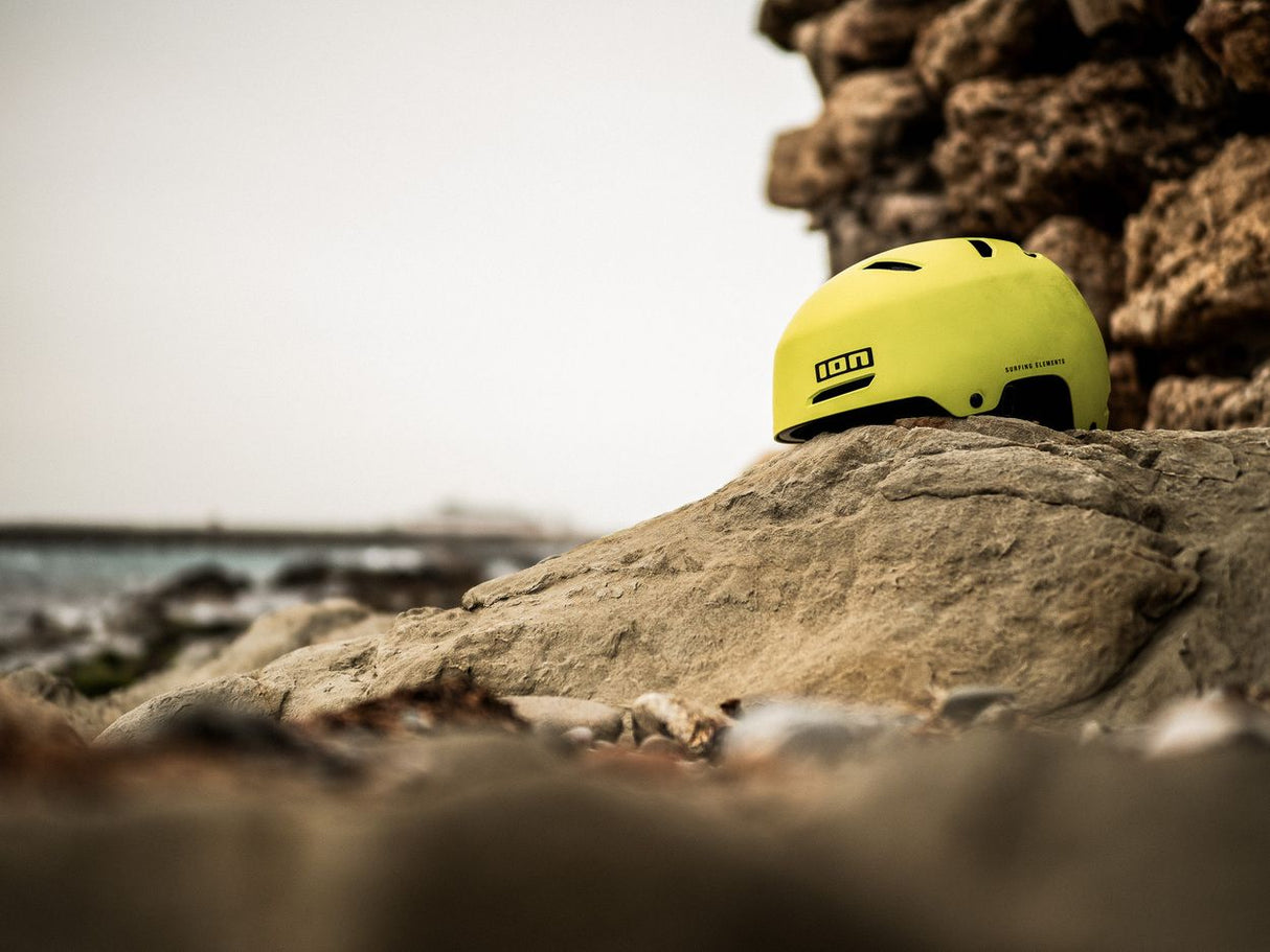 ION Slash Core Helm 2026 in Neongelb, Seitenansicht auf Stein am Strand, robuster Watersport-Helm mit Belüftungsöffnungen