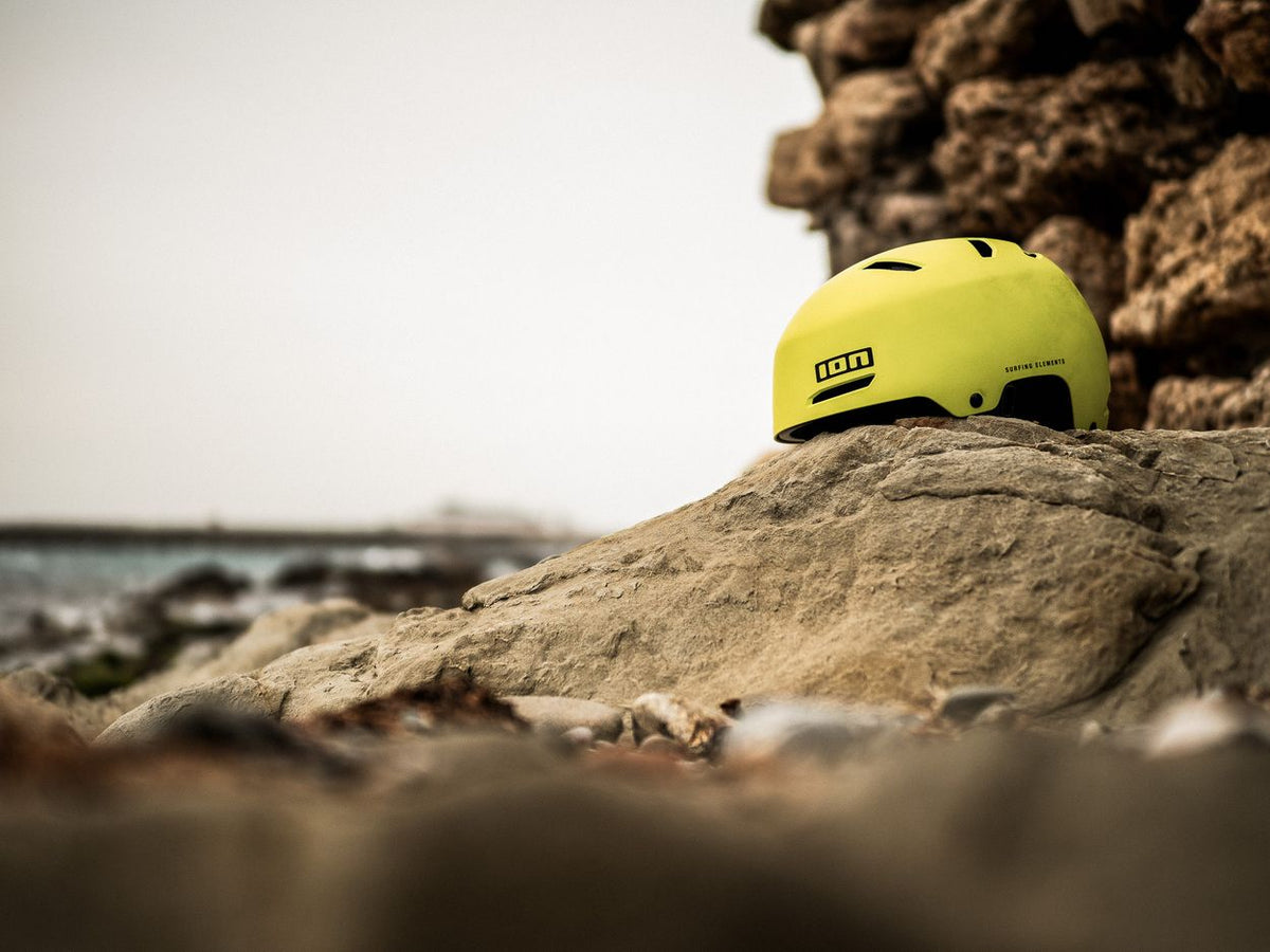 ION Slash Core Helm 2026 in Neongelb, Seitenansicht auf Stein am Strand, robuster Watersport-Helm mit Belüftungsöffnungen