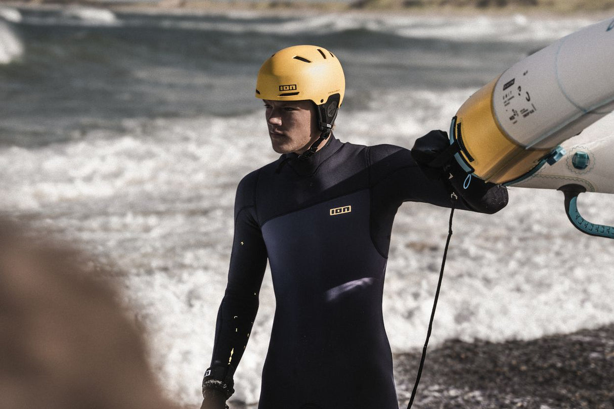ION Slash Amp 2026 Kiteboard Neoprenanzug schwarz, Sportler trägt gelmen Helm am Strand mit Board