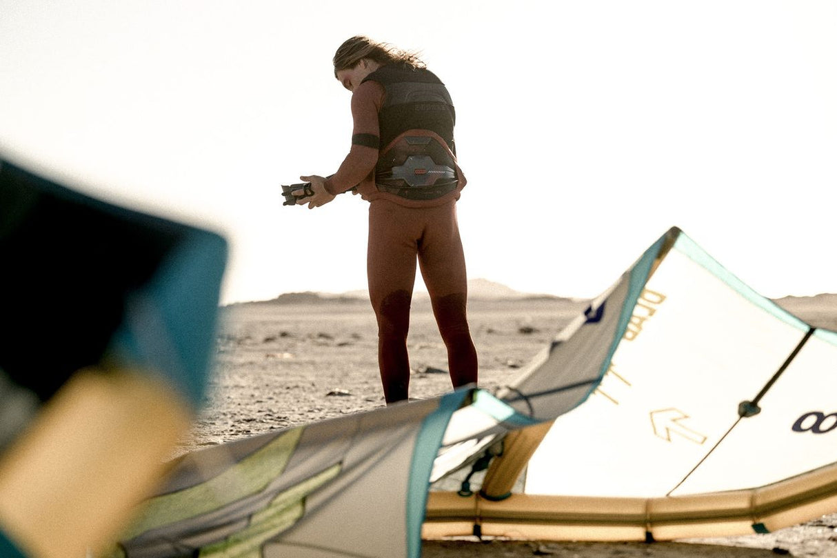 ION Apex Kite Trapez Herren 2026 in Braun, Athlet trägt Trapez am Strand mit Kite