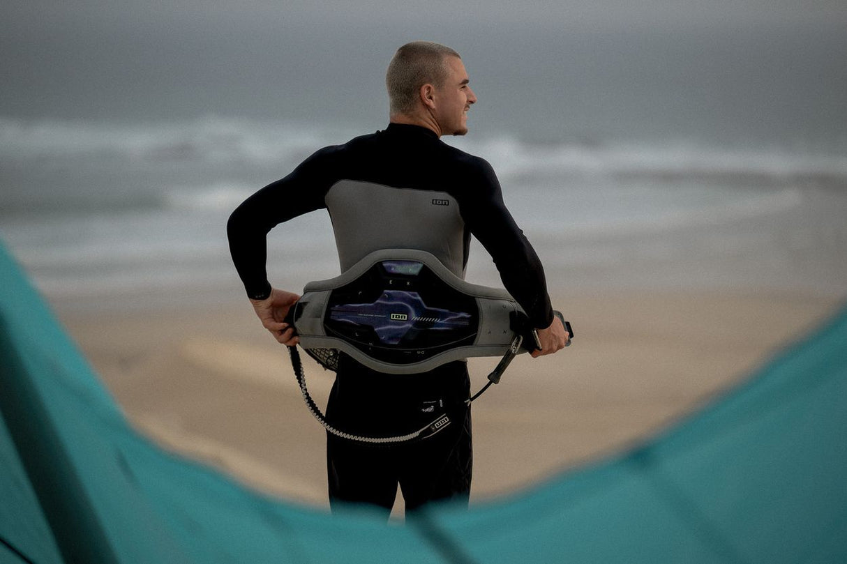 ION Apex Kite Trapez Herren 2026 in Schwarz-Grau, von vorne getragen am Strand, mit Kite im Hintergrund