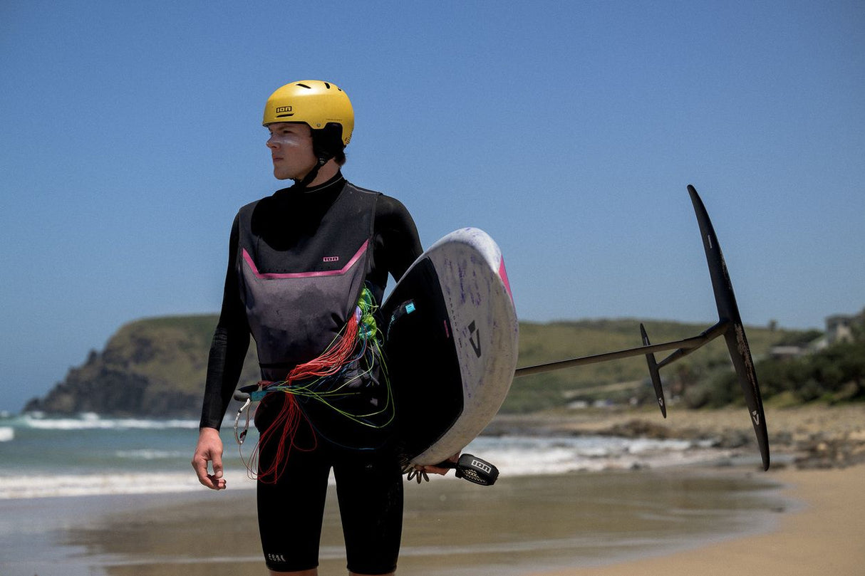 ION Neo Jacket Jam schwarze Neopren-Jacke, Frontalansicht am Strand mit Kiteboard und gelbem Helm