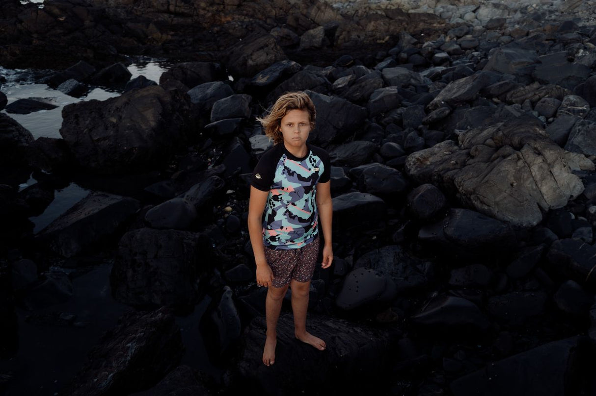 ION Rashguard Capture 2026 in Schwarz mit Muster, Kurzarm, getragen an Felsen am Strand