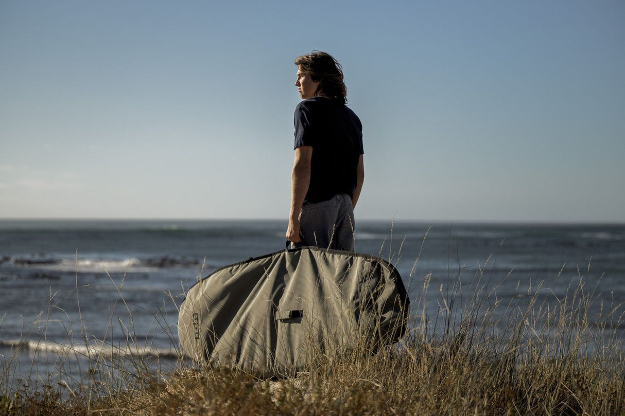 ION Wing Boardbag Core 2026 Transporttasche in Beige, Mann trägt Boardbag am Strand mit Meer im Hintergrund