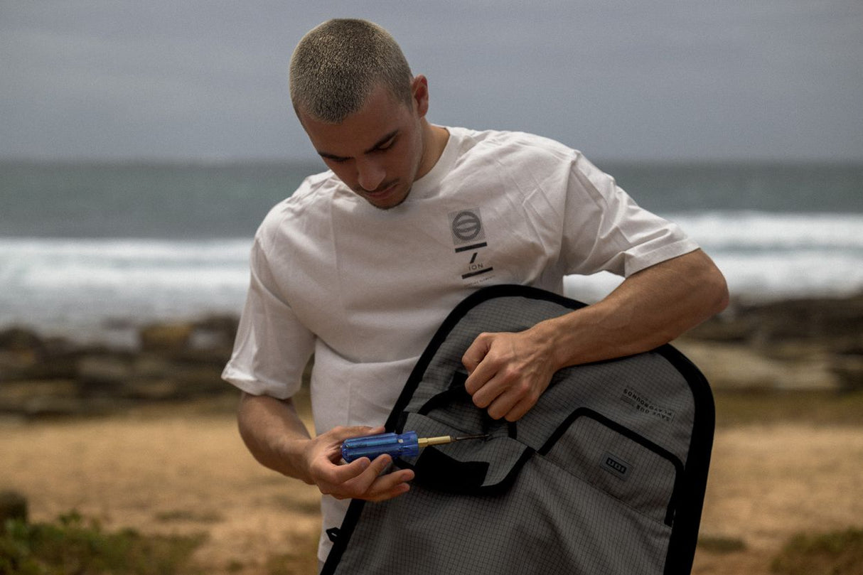ION Twintip Boardbag Core 2024 schwarz, Mann öffnet Tasche am Strand, Frontalansicht