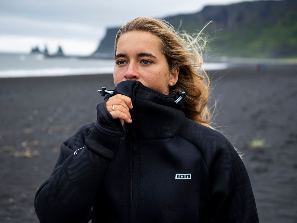 ION Neo Shelter Jacket 2024 schwarz, Frau trägt Jacke mit hochgezogenem Kragen am Strand, Frontalansicht