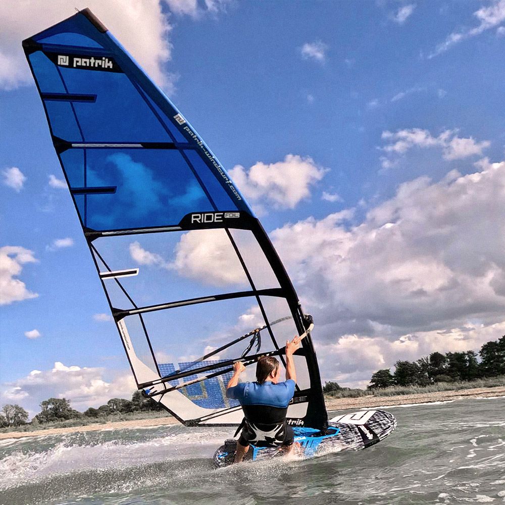 Patrik Foil Ride Windsurfboard blau mit Segel, Fahrer im Wasser beim Foilen, Seitenansicht