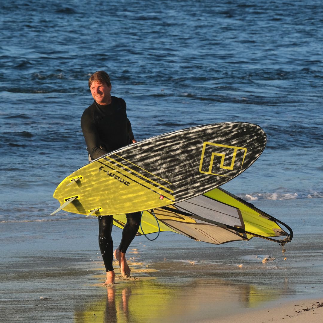 Patrik F-Wave Wellenreiter mit gelbem und schwarzem Surfboard am Strand mit Welle im Hintergrund