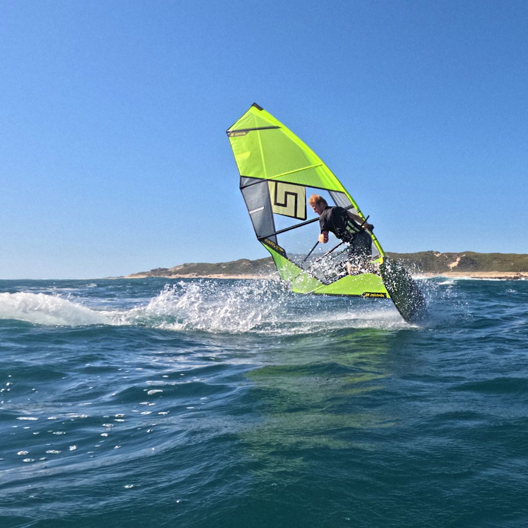 Patrik F-Wave Windsurfboard mit neongelber und grauer Segel beim Wellenreiten im Meer, Seitenansicht