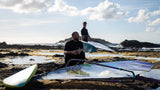 Duotone Super_Hero D/LAB 2027 Kiteboard in Blau und Weiß, am Strand mit Kitesurfern, Frontalansicht