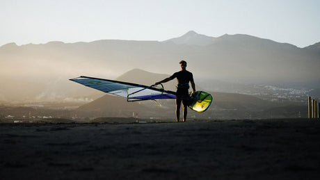 Duotone Sail Idol 2027 Windsegel in Blau und Grün, Sportler in Neoprenanzug hält Segel am Strand vor Bergkulisse
