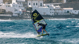 Duotone Sail Idol 2027 Windsurfboard mit schwarzem und gelbem Segel, Fahrer auf Welle in blauem Meer, Küstensiedlung im Hintergrund