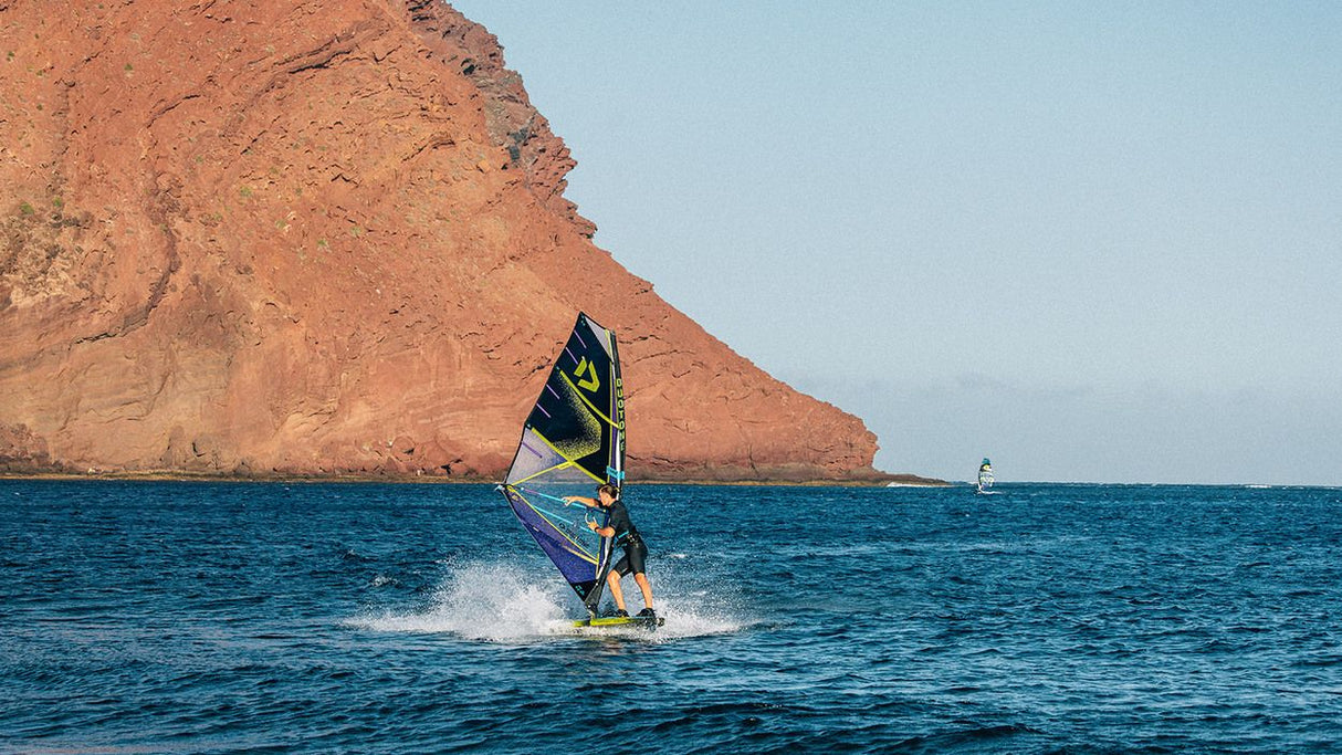 Duotone Sail Idol 2027 Windsegel in Gelb und Schwarz, Windsurfer auf dem Wasser vor Felsenküste