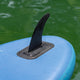 Ariinui Mahana SUP-Finne schwarz auf blauem Board, Detailansicht der gebogenen Finne mit Befestigungsbox im Wasser
