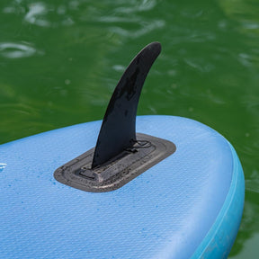 Ariinui Mahana SUP-Finne schwarz auf blauem Board, Detailansicht der gebogenen Finne mit Befestigungsbox im Wasser