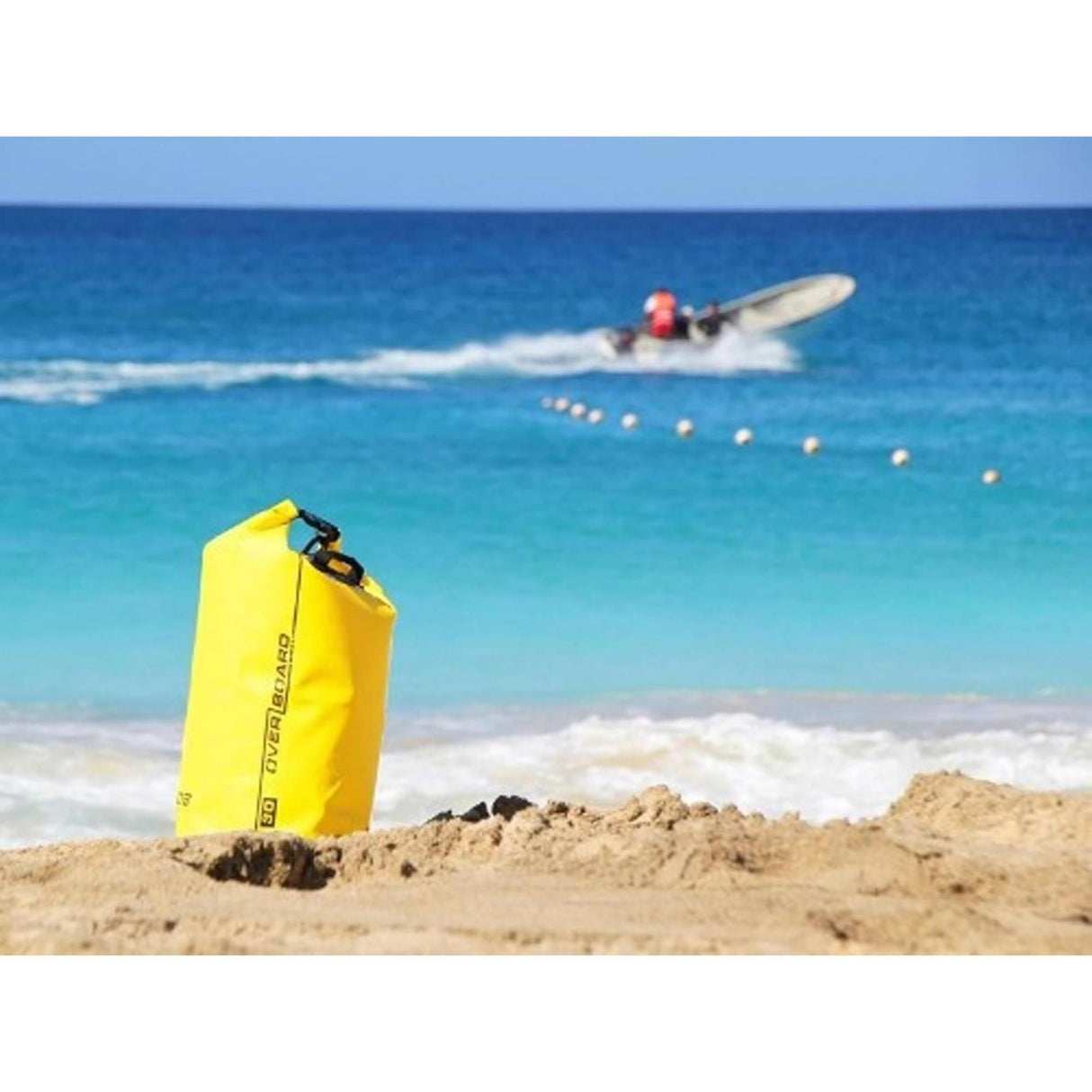 OverBoard wasserdichter Packsack 30 Liter gelb am Strand, im Hintergrund Wassersportler mit Jetski