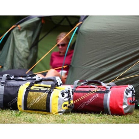 OverBoard Duffel Bag Pro wasserdichte Reisetasche in Schwarz, Gelb und Rot, liegend auf Gras vor Zelten