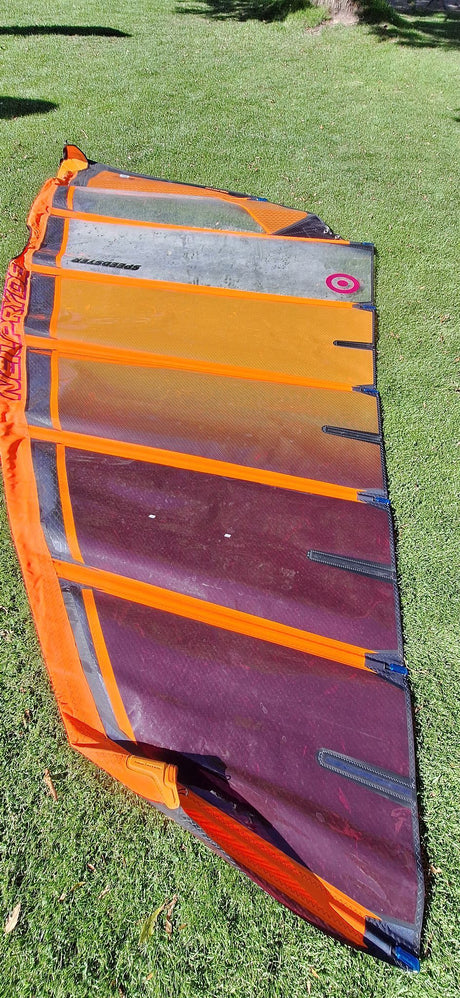 Orange and purple windsurfing sail laid out on green grass, showing full design and structure with visible brand text and seams.