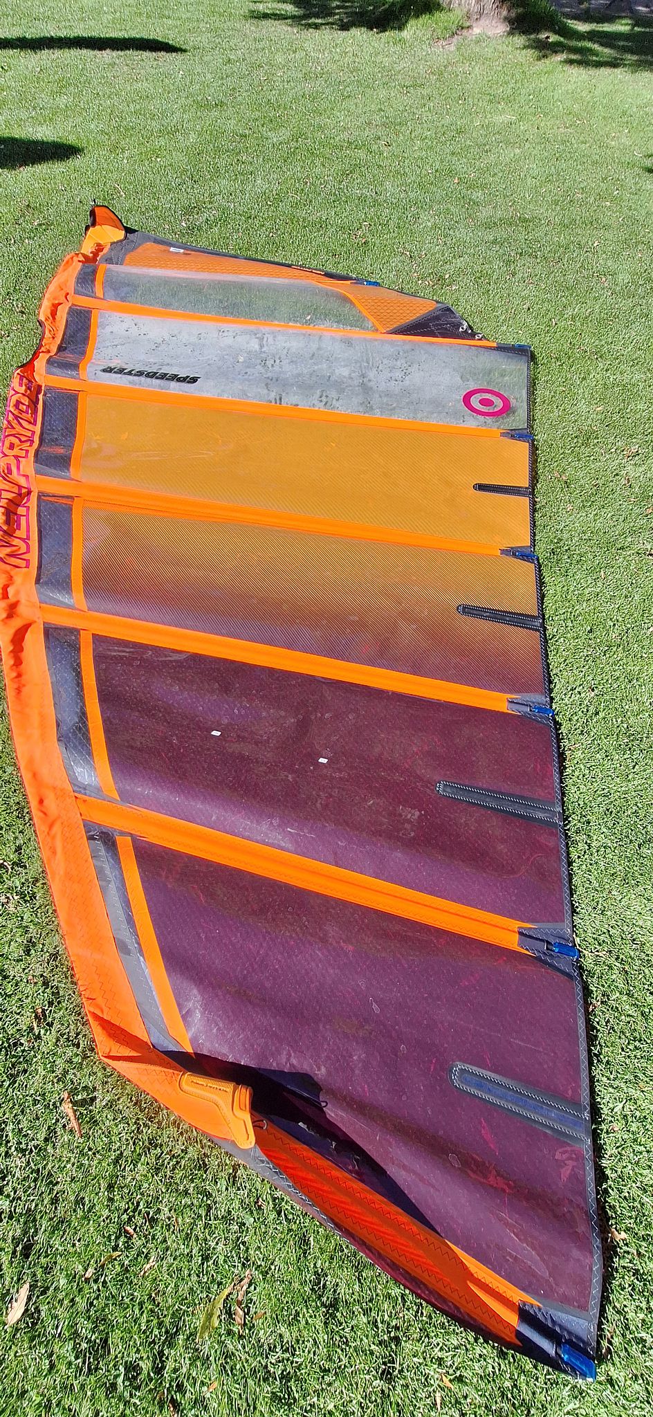 Orange and purple windsurfing sail laid out on green grass, showing full design and structure with visible brand text and seams.