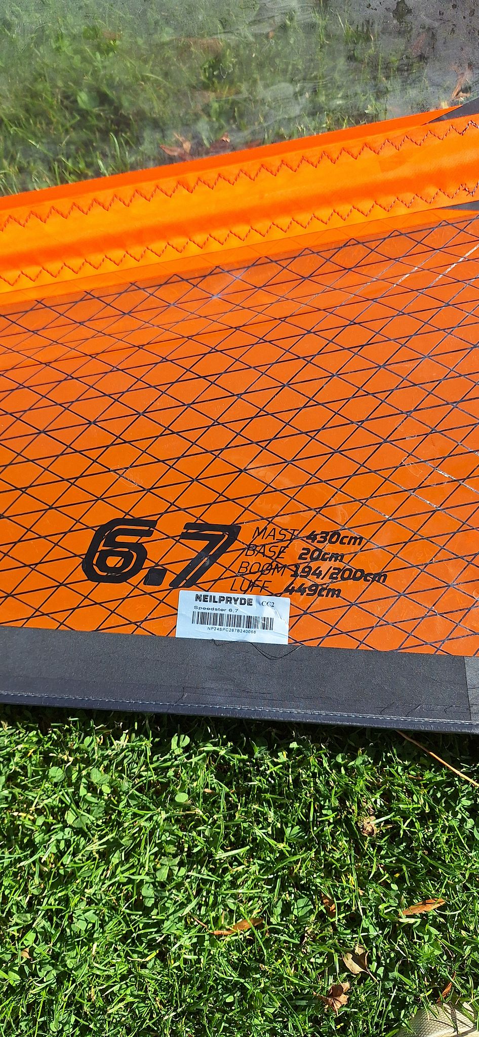 Close-up of a NeilPryde 6.7 windsurfing sail with orange diamond-patterned fabric, showing specifications and branding, resting on grass.
