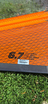 Close-up of a NeilPryde 6.7 windsurfing sail with orange diamond-patterned fabric, showing specifications and branding, resting on grass.