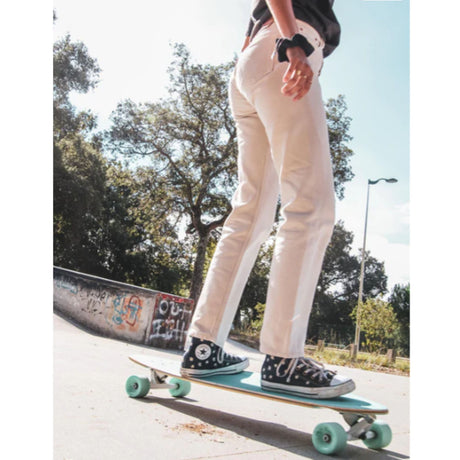 Flying Wheels Praise Gun Skateboard mit grünen Rollen, Person in weißer Hose beim Skaten im Park, Frontalansicht
