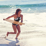 Skimboard SkimOne Palm Beach in Holzoptik, von einer Frau am Strand getragen, Meerwasser im Hintergrund