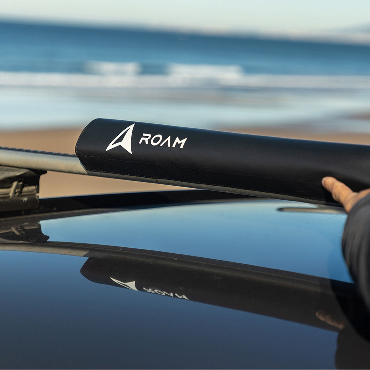 ROAM Dachauflage Surfboard Rund, schwarze Schutzhülle mit ROAM-Logo auf Autodach, Strandkulisse im Hintergrund