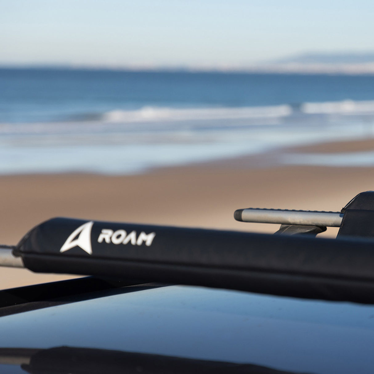 ROAM Dachauflage Surfboard Flach schwarz auf Autodach, Strandszene im Hintergrund