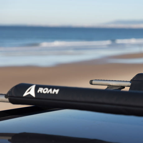 ROAM Dachauflage Surfboard Flach schwarz auf Autodach, Strandszene im Hintergrund