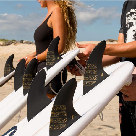 Futures Thruster Fin Set Vector 3/2 Alpha Medium, schwarze Flossen mit goldenem Muster auf Surfbrett, Strandansicht