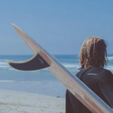 Futures Single Fin Albacore Flex Fiberglass, beige-rosé, Surfer trägt Flosse am Strand mit Meeresblick